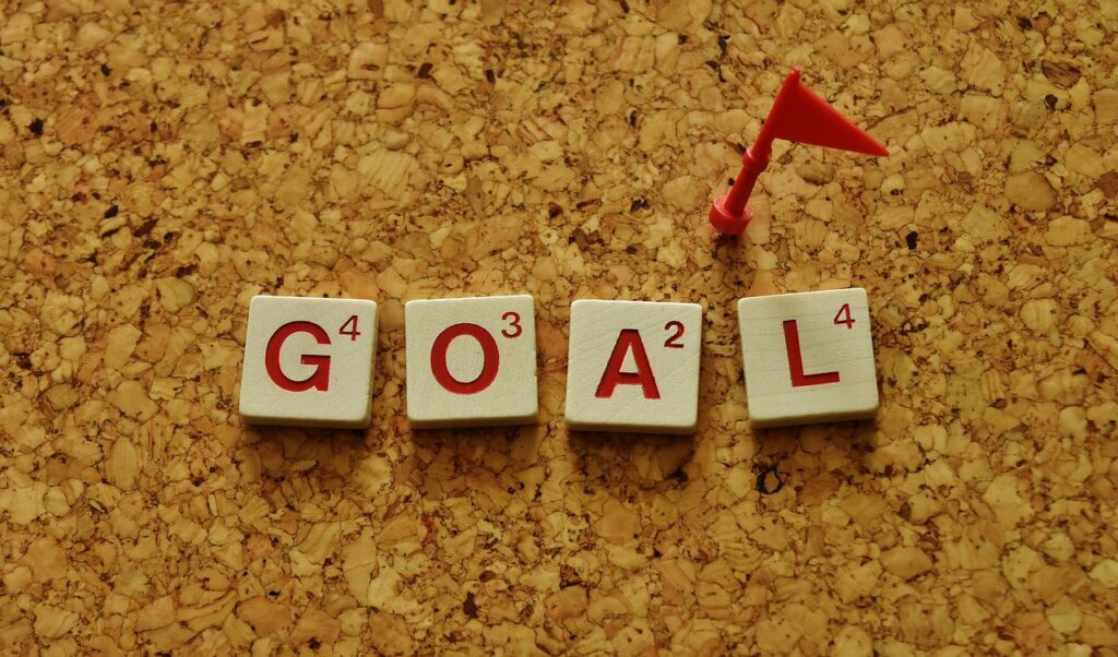 The word "goal" is spelled out on small wooden square block pieces, with the letters being in a vivid red. They are sat on top of a corkboard, with a small red plastic flag pinned above them. 