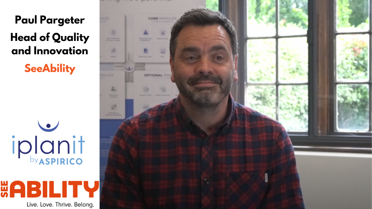 A man in a red and black checkered shirt sits indoors with a 